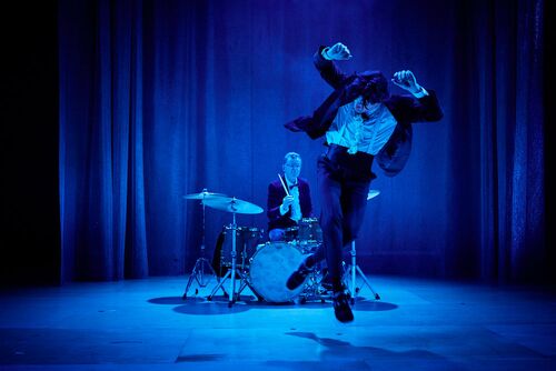 Conor Guilfoyle at a drumset with Alex O'Neill in the air in movement in front of him